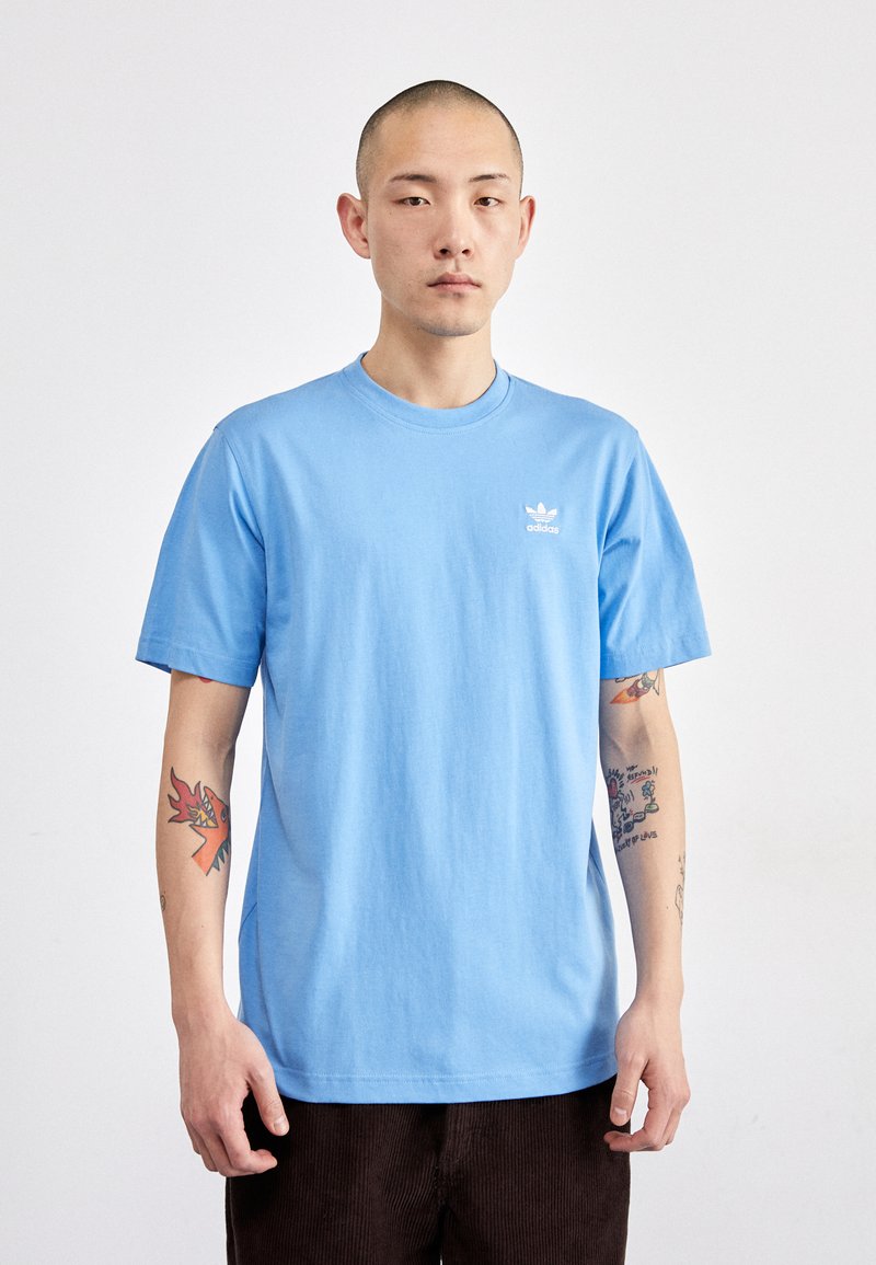 Camiseta de algodón de color azul claro con cuello redondo y mangas cortas. Presenta un pequeño logo blanco de Adidas en el lado izquierdo del pecho. Diseño simple.