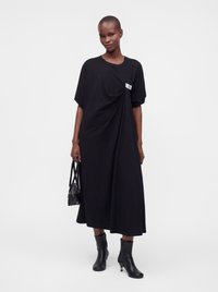 Femme mince portant une robe midi noire ample, tenant un sac à main noir et portant des bottines noires, debout devant un fond uni.
