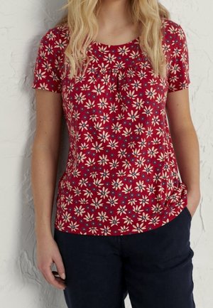Vrouw draagt een rood shirt met korte mouwen met wit en blauw bloemmotief en donkere broek, staand tegen een gestructureerde witte muur.