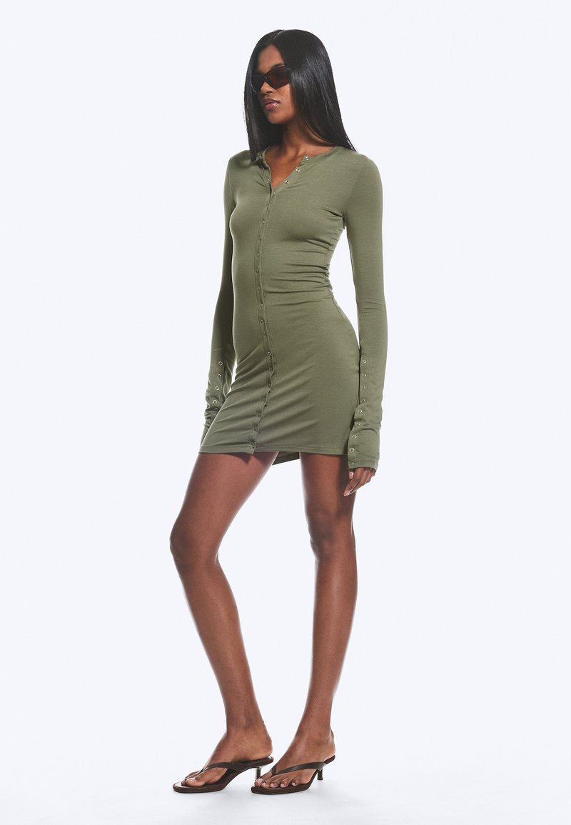 Femme portant une mini-robe à manches longues olive avec boutons sur le devant, lunettes de soleil foncées et sandales à talons noires, debout devant un fond blanc.