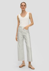 Weißes ärmelloses Oberteil mit V-Ausschnitt, kombiniert mit hochgeschnittenen, weit geschnittenen hellgrauen Jeans. Das Model trägt beige Zehentrenner.