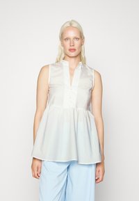 Blusa sem mangas de cor clara com colarinho alto, botões na frente e bainha franzida e rodada. Tecido suave e ligeiramente brilhante.