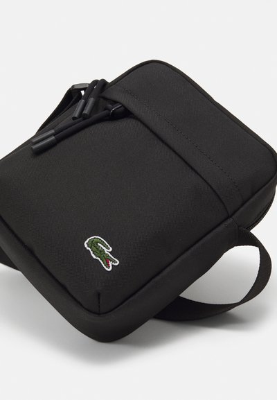 Sac à épaule en tissu noir avec fermeture éclair, présentant un petit logo de crocodile vert brodé et une finition texturée.