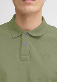 Polo vert olive en tissu texturé, doté d'un col classique, de deux boutons bleus et d'une discrète broderie de logo sur la poitrine.