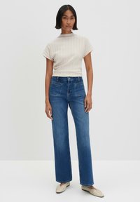 Beige geribbeld top met korte mouwen, gecombineerd met wijde blauwe jeans. Het model draagt lichtgekleurde flats en staat tegen een witte achtergrond.