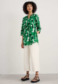 Groene, gemarmerde tuniek met abstracte vormen, losse pasvorm, driekwartmouwen, gecombineerd met witte wijde broek en zwarte sandaaltjes met bandjes.