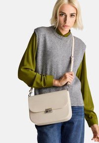 Borsa a tracolla beige con texture, dotata di patta e hardware argentato. Indossata da una persona con un maglione grigio senza maniche e una camicia verde a maniche lunghe.