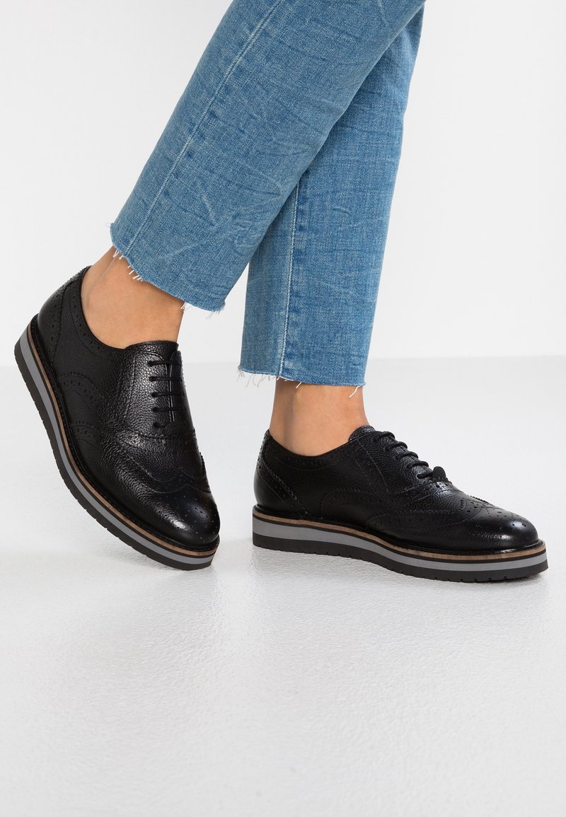 Pieds portant des chaussures brogue en cuir noir avec des semelles épaisses rayées et un jean bleu court effiloché sur un fond de sol clair.