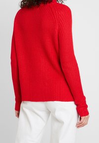 Pull en maille rouge avec une coupe décontractée, texture côtelée et manches longues. Associé à un pantalon blanc, mettant en valeur un look décontracté superposé.
