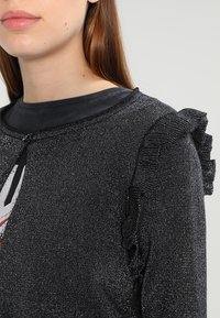 Maglione in maglia metallici nera con scollo rotondo, dettagli arricciati sulle spalle e un colletto nero liscio.