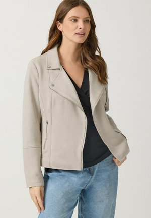 Frau trägt eine beige Reißverschlussjacke über einem schwarzen Oberteil und blauen Jeans, steht vor schlichtem hellem Hintergrund, mit einer Hand in der Tasche.