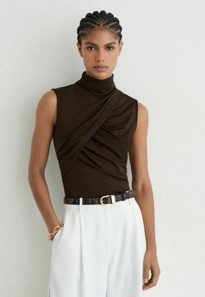 Frau mit geflochtenem Haar, die ein ärmelloses schwarzes Rollkragentop, weiße Hose, einen dunklen Gürtel und ein goldenes Armband trägt, steht vor einer weißen Wand.