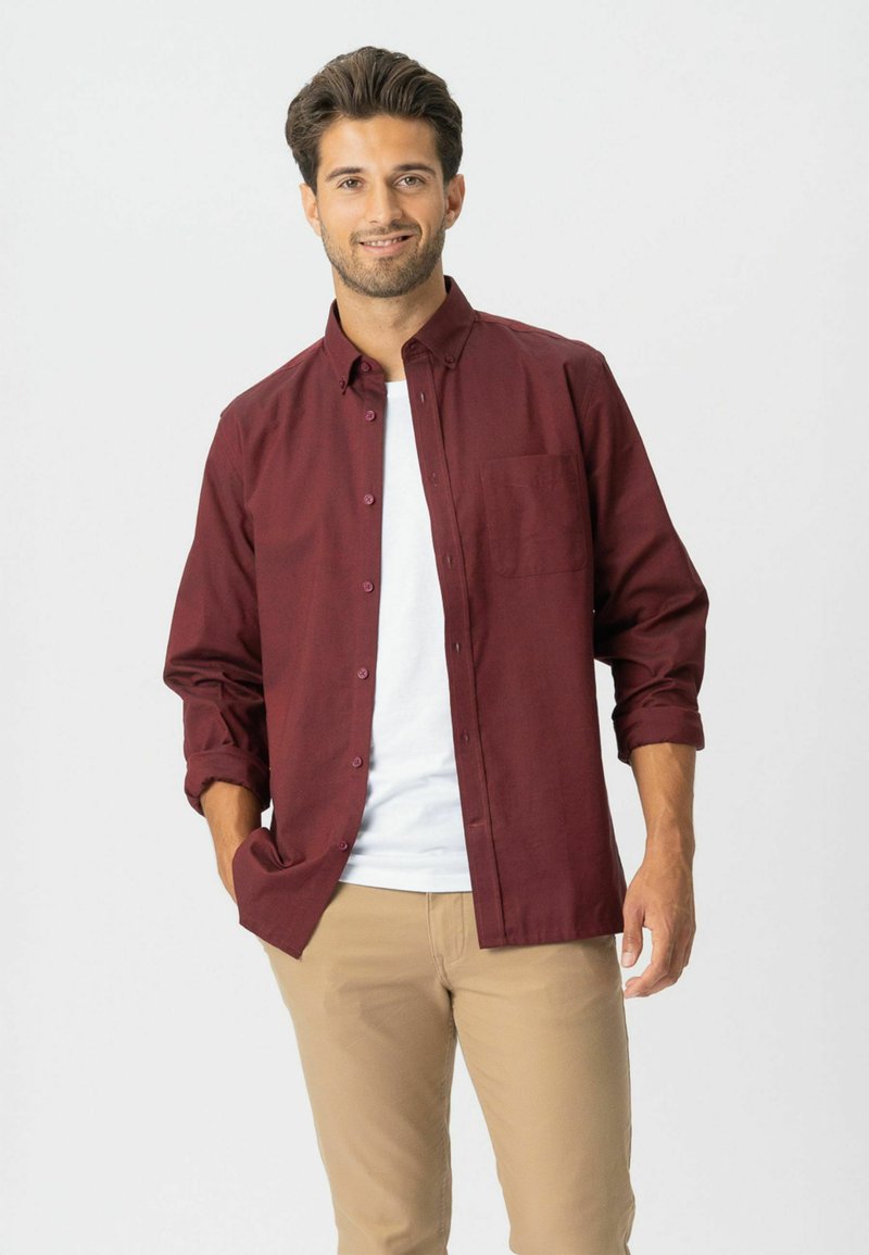 Hombre con cabello oscuro que lleva una camisa de botones color burdeos sobre una camiseta blanca y pantalones beige, de pie con una mano en el bolsillo, sonriendo.