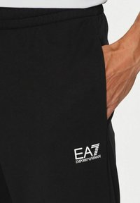 Pantaloni della tuta neri realizzati in tessuto misto cotone, dotati di vita elastica, tasche laterali e logo bianco EA7 Emporio Armani sulla coscia.
