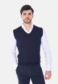 Gilet senza maniche lavorato a maglia di colore blu navy con scollatura a V, indossato sopra una camicia bianca con bottoni, abbinato a pantaloni scuri. Tessuto morbido, design semplice.