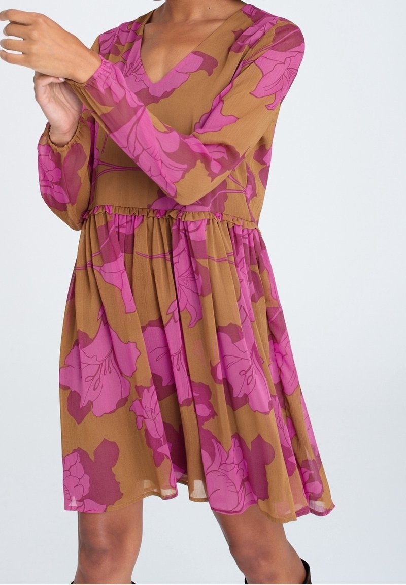 Femme portant une robe marron avec de grandes imprimés floraux roses, manches longues, col en V, et taille froncée, vue des épaules aux genoux.