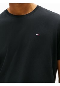 Camiseta negra de manga corta con cuello redondo y un pequeño logo bordado en el pecho con franjas rojas, blancas y azules.