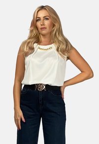 Biała bezrękawnik z złotym łańcuszkiem przy dekolcie, zestawiony z ciemnoniebieskimi spodniami z wysokim stanem i czarnym pasem z złotą klamrą.