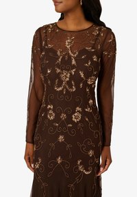 Hnědé večerní šaty s dlouhými průsvitnými rukávy, zdobené sofistikovanou zlatou květinovou výšivkou a korálkovými akcenty na dobře padnoucím siluetě.
