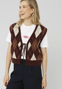 Gilet argyle marron et crème avec un col en V et un système de fermeture par lien, porté par-dessus un T-shirt blanc avec un texte rouge. Un pantalon à rayures noires vient compléter la tenue.