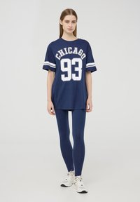 T-shirt oversize blu navy con grafica "CHICAGO 93", strisce bianche sulle maniche, abbinato a leggins blu navy e scarpe sportive bianche.