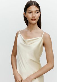 Ärmlös klänning i satin med cowl-halsringning i krämfärg, med en mjuk textur och avslappnad silhuett. Modellen bär ett delikat halsband i guld.