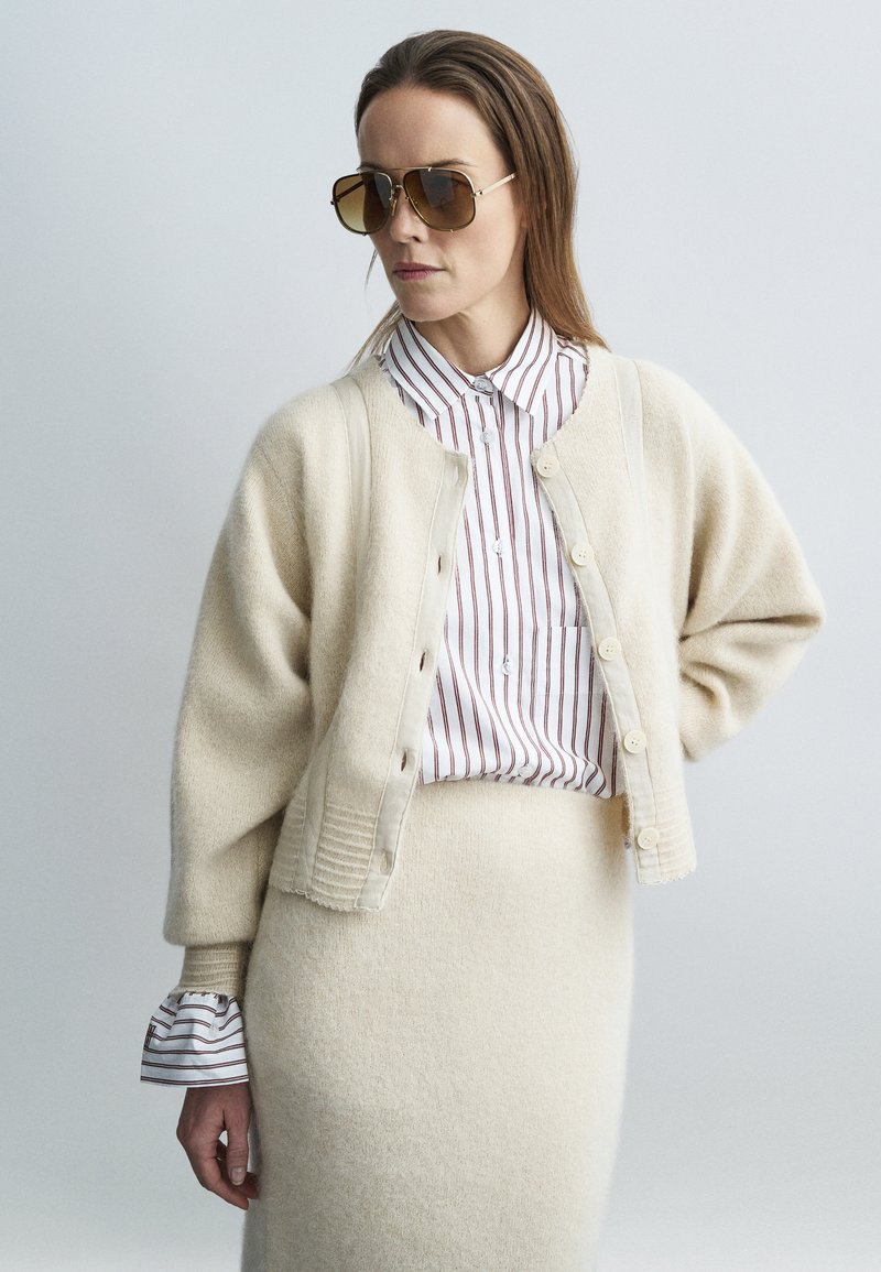 Beež kardigan nööpidega, pehme tekstuuriga, kombineeritud triibulise särgi ja sobiva seelikuga. Üle suurusega päikeseprillid rõhutavad stiili.