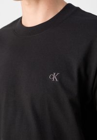 Czarna bawełniana koszulka z okrągłym dekoltem, z małym szarym haftowanym logo "cK" po lewej stronie klatki piersiowej. Gładka tekstura.