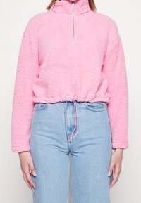 Pullover in pile rosa con colletto alto, zip frontale e orlo elasticizzato. Presenta un tessuto texturizzato e dettaglio con coulisse in vita.