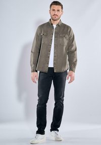 Olivgrünes, knöpfbares Hemd mit zwei Brusttaschen, getragen über einem weißen T-Shirt, kombiniert mit schwarzen Jeans und weißen Sneakern.