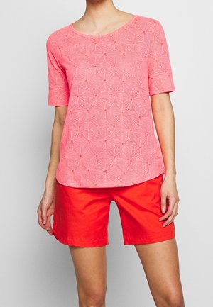 Femme portant un haut à manches courtes rose avec un motif et un col arrondi, ainsi qu'un short rouge vif, debout devant un fond clair.