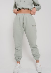 Mintgrüne Sweatpants mit einem elastischen Taillenbund und Bündchen, ausgestattet mit einem Kordelzug und einem prägten Logo an der Seite, kombiniert mit weißen Turnschuhen.