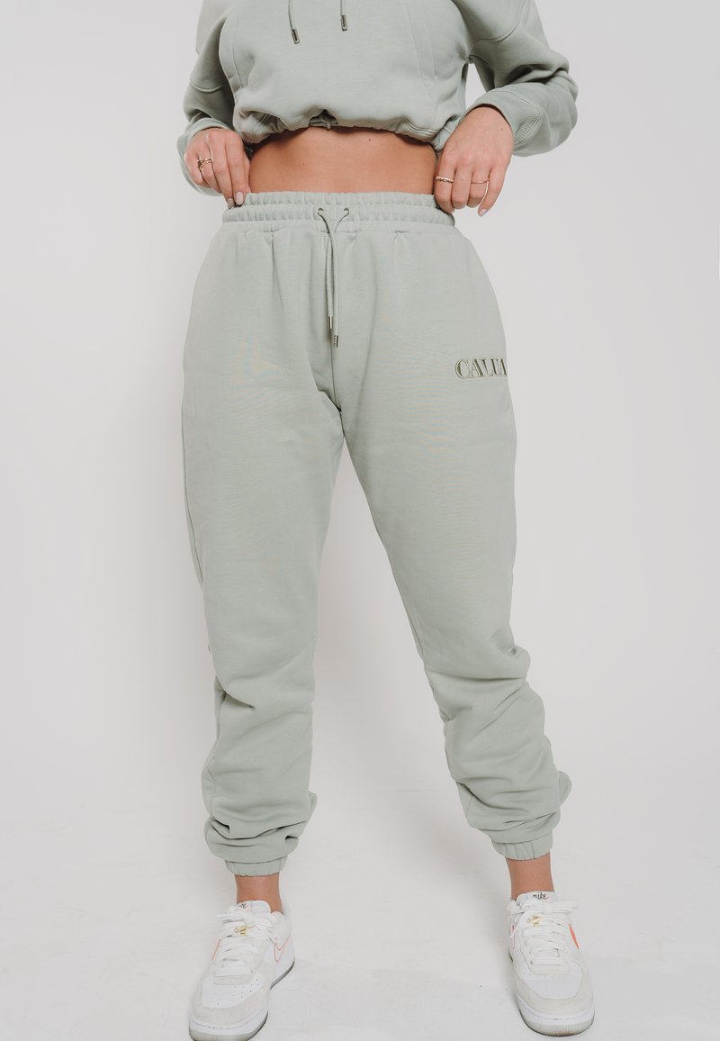 Mintgrüne Sweatpants mit einem elastischen Taillenbund und Bündchen, ausgestattet mit einem Kordelzug und einem prägten Logo an der Seite, kombiniert mit weißen Turnschuhen.