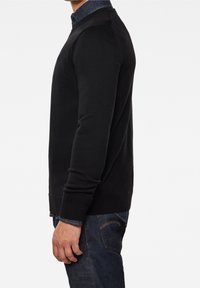 Schwarzer Strickpullover mit Rundhalsausschnitt und gerippten Bündchen, getragen über einem blauen Hemd mit Kragen, kombiniert mit dunkelblauen Jeans.