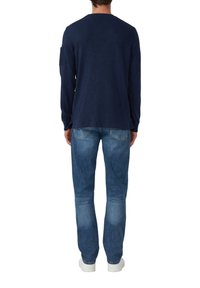 Man die met zijn rug naar de camera staat, gekleed in een marineblauwe longsleeve shirt, blauwe jeans met achterzakken en witte schoenen, op een witte achtergrond.