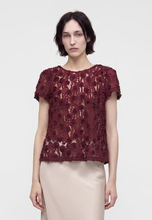 Femme aux cheveux courts et foncés portant un chemisier bordeaux transparent avec des appliques florales texturées et une jupe beige, debout sur un fond uni.