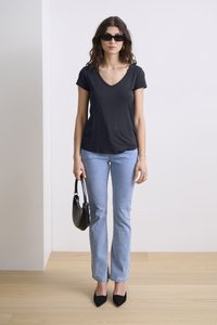 Fekete V nyakú póló, világoskék egyenes szárú farmer, fekete hegyes orrú magassarkú cipő és fekete kézitáska. A modellen sötét napszemüveg van.