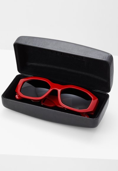Ochelari de soare roșii cu un design geometric îndrăzneț, în interiorul unei carcase din plastic rigid negru, având lentile întunecate și branding vizibil pe carcasă.