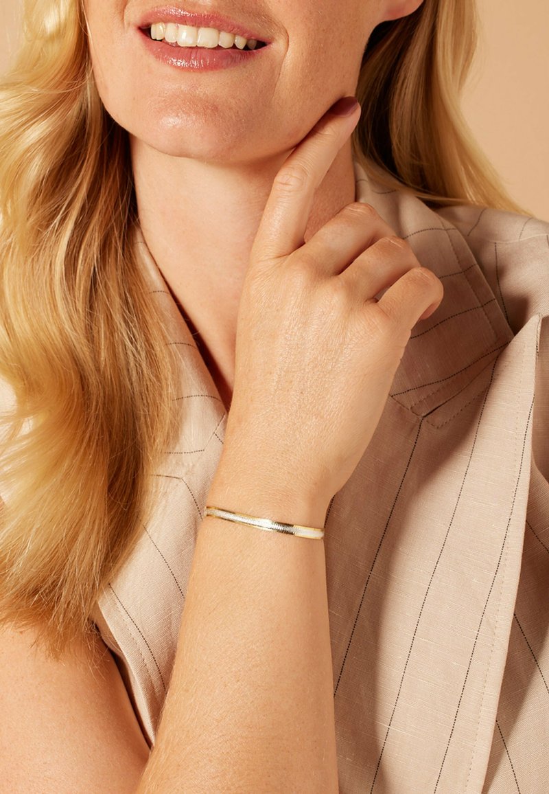 Gold-tone metal bracelet with a smooth finish, featuring a slight texture and a slender design. Worn on a wrist, paired with a light fabric top.