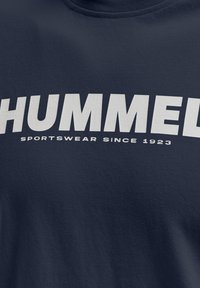 Tumši zila T-krekls ar uzkrītošiem baltiem burtiem, uz kuriem rakstīts "HUMMEL SPORTSWEAR SINCE 1923", izgatavots no mīksta kokvilnas auduma.