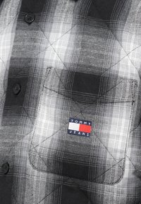 Fekete-fehér kockás gombos ing, mellzsebbel, amelyen egy színes címke látható "TOMMY JEANS" felirattal. Az anyag texturált felületű.