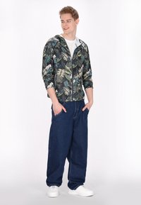 Felpa con zip floreale in tonalità scure con foglie verdi e bianche, abbinata a pantaloni di denim blu scuro di taglio largo e sneakers bianche.