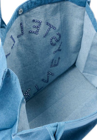 Vista interna di una borsa tote in denim azzurro chiaro con lettere di perline blu scuro disposte in un motivo circolare sul tessuto interno.