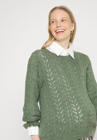 Suéter tricotado verde claro com um padrão texturizado de folhas, gola canelada e mangas bufantes, com uma camisa branca por baixo.