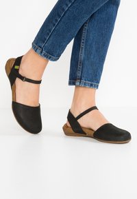 Černé kožené slingback boty s kulatou špičkou a kotníkovým páskem. Koreksole s texturovaným povrchem, spárované s tmavými cropped džínami.
