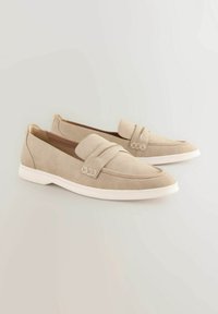 Beige suede loafers with a slightly raised heel, featuring a textured toe cap and a decorative strap across the vamp with metal accents.