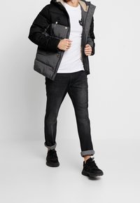 Giacca a vento imbottita in nero e grigio, con cappuccio foderato in pelliccia, bottoni a pressione e logo sul petto. Abbinata a jeans scuri e sneakers nere.