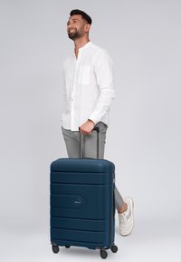 Uomo con la barba che indossa una camicia bianca e pantaloni grigi, in piedi, sorridente, mentre tiene il manico di una grande valigia blu con le ruote.