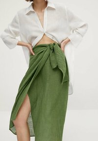 Chemise blanche à boutons, avec une coupe décontractée, associée à une jupe portefeuille en lin vert taille haute, dotée d'un nœud et d'une fente sur le côté.