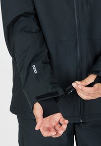 Svart utomhusjacka med hög krage, dragkedjor på fickorna, justerbara kardborremuddar och en texturerad tyg med vattentåliga detaljer.